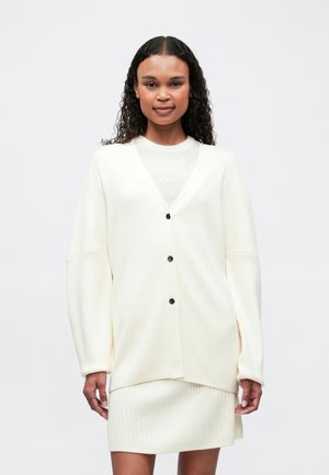 Une femme aux cheveux bouclés porte un cardigan blanc à boutons sur une jupe blanche côtelée, se tenant devant un fond léger uni.