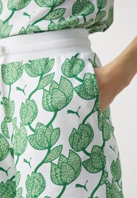 Valkoiset shortsit, joissa on vihreä kukkakuvio ja pieniä Puma-logoja, näyttäen käden tunkeutuvan sivutaskuun.