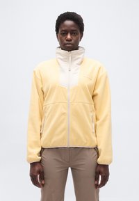 Άτομο που στέκεται με πρόσωπο προς τα εμπρός, φοράει μια ζιπ-λεπτή φλις μπουφάν σε απαλό κίτρινο χρώμα με μπεζ πινελιές και ανοιχτό καφέ παντελόνι, μπροστά σε απλο υπόβαθρο.