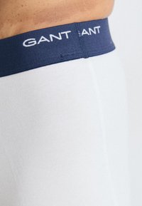 Gros plan d'une personne portant un sous-vêtement blanc avec une ceinture bleu marine sur laquelle est inscrit le nom de la marque "GANT" en lettres blanches.