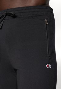 Zwarte sportbroek met een koord in de taille, voorzien van een ritszak en een klein geborduurd logo op de dij. Soepele textuur.