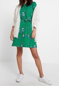 Zelené květinové šaty se zavazováním v pase, krátkými rukávy a splývavým lemem, doplněné texturovaným bílým kardiganem a bílými teniskami.