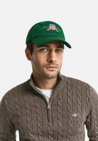 Grüne Mütze mit aufgesticktem Logo in Rot, Weiß und Blau; kombiniert mit einem braunen Strickpullover und einem weißen Hemd darunter.