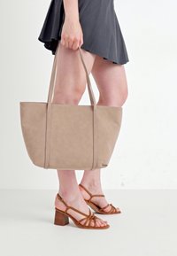 Beige Tragetasche mit doppelten Griffen, aus glattem Material gefertigt, mit strukturierter Form und minimalistischem Design. Dazu werden braune Riemchen-Heels getragen.