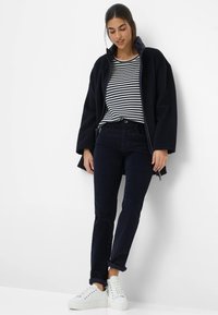 Vrouw die tegen een witte muur staat, gekleed in een zwarte jas, een zwart-wit gestreept shirt, donkere broek en witte sneakers, met een ontspannen houding naar beneden kijkend.