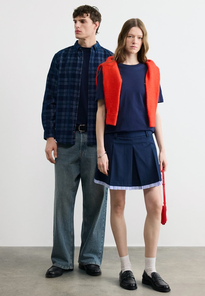 Ένας άνδρας με μπλε καρό πουκάμισο και φαρδιά τζιν στέκεται δίπλα σε μια γυναίκα με μπλε φούστα, μπλε πουκάμισο και κόκκινο πουλόβερ ριγμένο στους ώμους, κρατώντας μια κόκκινη τσάντα.