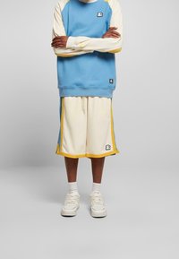 Persona che indossa una felpa blu e crema con pantaloni corti crema abbinati con bordi gialli, calzini bianchi e sneakers bianche, in piedi con le braccia incrociate.