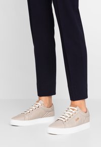 Pieds portant des baskets basses beiges avec des lacets et semelles blanches, associés à un pantalon bleu marine ajusté sur un fond uni.