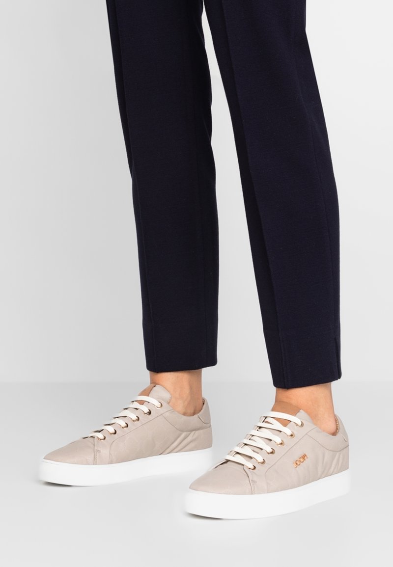 Pieds portant des baskets basses beiges avec des lacets et semelles blanches, associés à un pantalon bleu marine ajusté sur un fond uni.