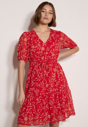 Młoda kobieta o krótkich brązowych włosach, ubrana w czerwoną sukienkę z beżowymi wzorami liści i bufkami na krótkich rękawach, stojąca na jednolitym tle.