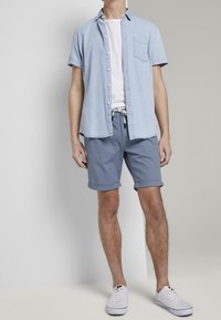 Camisa azul clara de manga corta con botones sobre una camiseta blanca, combinada con pantalones cortos grises enrollados y zapatillas blancas.