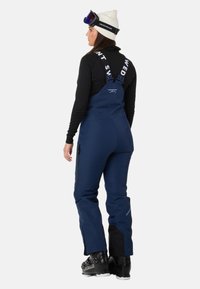 Navyblauwe skibroek met verstelbare banden, zijritszakken en versterkte onderbenen. Gecombineerd met zwarte skirchoenen en een witte beanie.