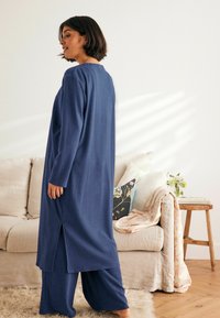 Tamsiai mėlynos spalvos laisvalaikio rinkinys, kuriame yra ilgas marškinėlis su šoninėmis plyšiais ir plačiomis kelnėmis, pagamintas iš minkštos, tekstūruotos medžiagos. Paprastas dizainas, laisvas siluetas.