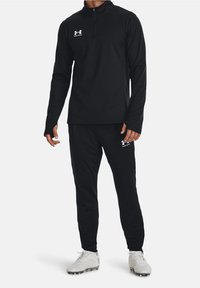 Schwarzer sportlicher Pullover mit Viertelreißverschluss und Logo auf der Brust, kombiniert mit passenden schwarzen, zulaufenden Hosen. Beide aus einem glatten, atmungsaktiven Material gefertigt.