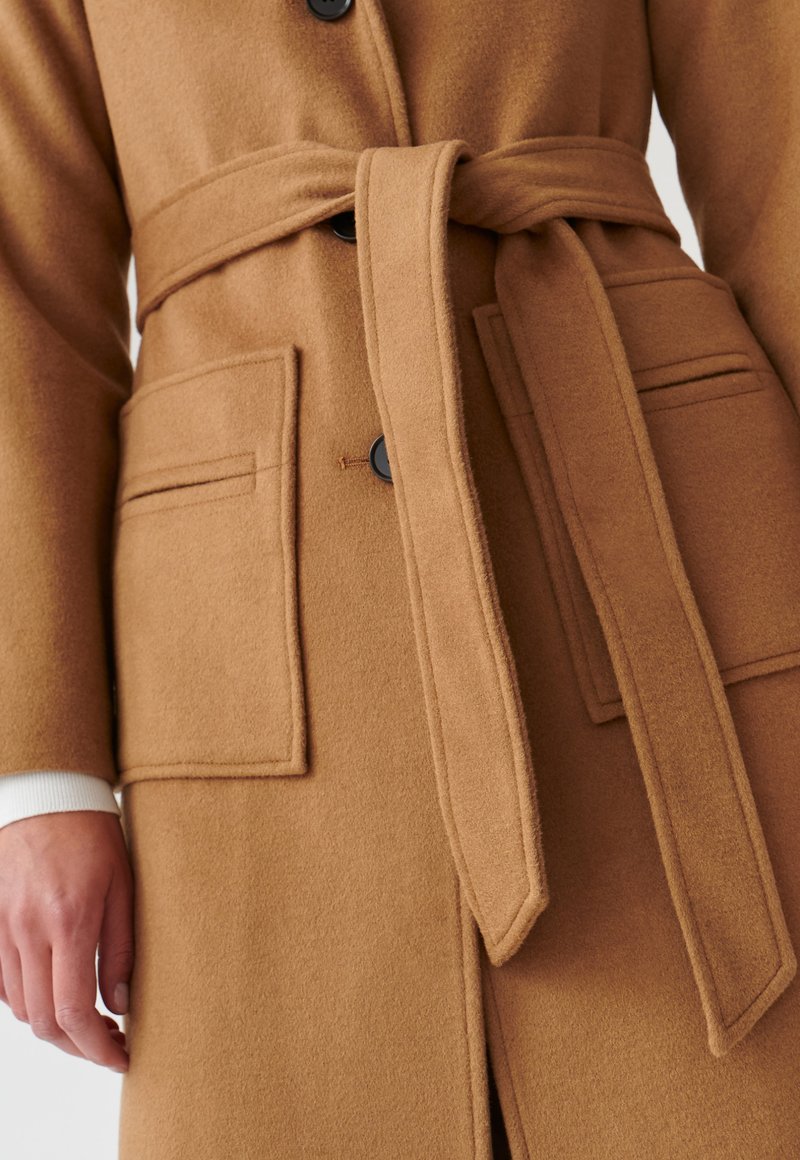 Cappotto in misto lana marrone con cintura legata, due tasche frontali, bottoni neri e una texture morbida, con un polsino bianco visibile sotto.