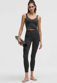Juodas treniruočių komplektas, sudarytas iš trumpąja tankiniu ir aukšto liemens leggingais su subtiliu gėlių raštu. Modelis laiko suvoliotą jogos kilimėlį.