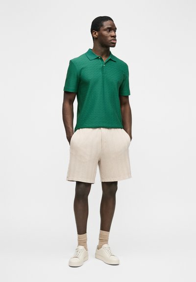 Άνδρας που φοράει πράσινο πόλο μπλουζάκι με κοντά μανίκια, μπεζ σορτς με υφή, μπεζ κάλτσες και λευκά αθλητικά παπούτσια, όρθιος με τα χέρια στις τσέπες.