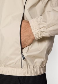 Bēšas krāsas viegla jaka ar elastīgām aprocēm un apakšmalu, kas ietver rāvējslēdzēja sānu kabatu un smalkus perforētus akcentus. Gluda tekstūra.