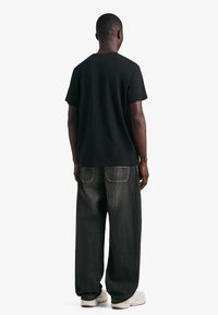 Tricou negru cu mâneci scurte, asortat cu blugi de denim închiși la culoare, largi. Are cusături albe și croială lejeră. Încălțămintea este compusă din adidași de culoare deschisă.