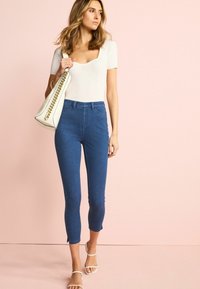 Vaqueros de mezclilla azules ajustados con cintura alta, de largo recortado y cortes laterales, combinados con una blusa blanca de canalé y sandalias de tiras beige.