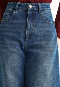 High-waisted blue denim jeans with a smooth texture, five pockets, and subtle stitching details. Button closure at the front.