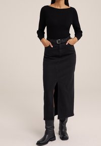 Haut noir à manches longues côtelées associé à une jupe en denim noire taille haute longueur cheville avec une fente à l'avant et une ceinture noire. Bottes noires montant jusqu'aux genoux.