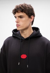 Felpa nera con cappuccio e coulisse, accentuata da un logo in gomma rossa sul petto e punte della coulisse in argento. Tessuto morbido e dall'aspetto texturizzato.