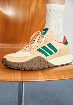 Baskets Adidas beige et vertes avec semelle marron texturée, portées avec une chaussette blanche côtelée, reposant sur une chaise en bois.