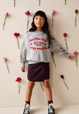 Pige står op ad en væg med tape-røde og pink roser, iført en grå sweatshirt med rød tekst, sort nederdel med hjerte-mønster og sorte støvler.