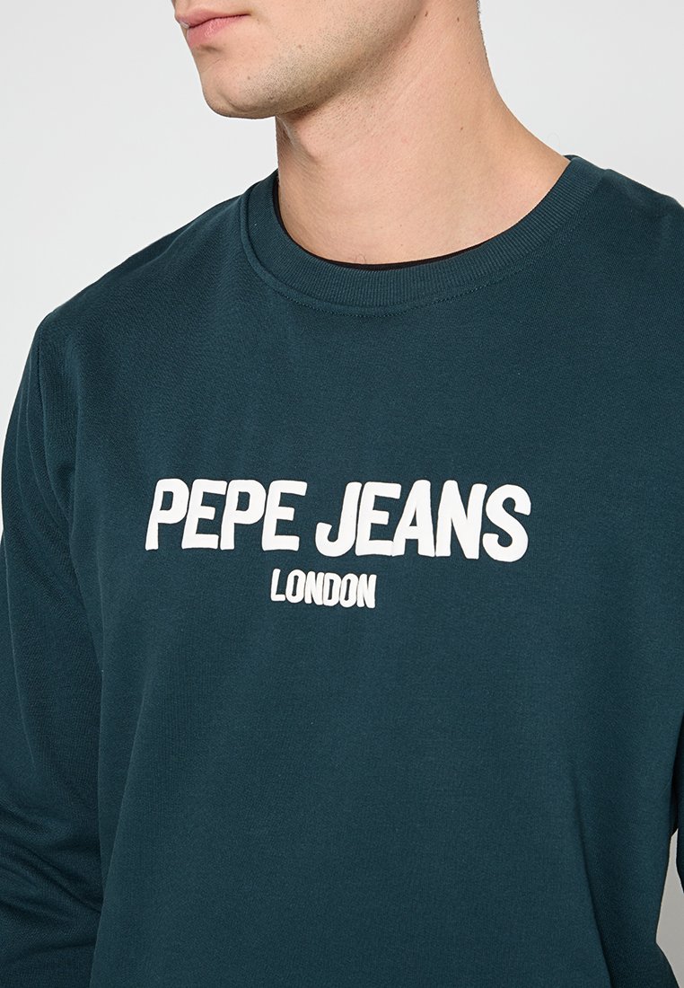 Camisa de manga larga color verde azulado, hecha de algodón, con un estampado blanco de "PEPE JEANS LONDON" centrado en el pecho. Cuello redondo, diseño simple.