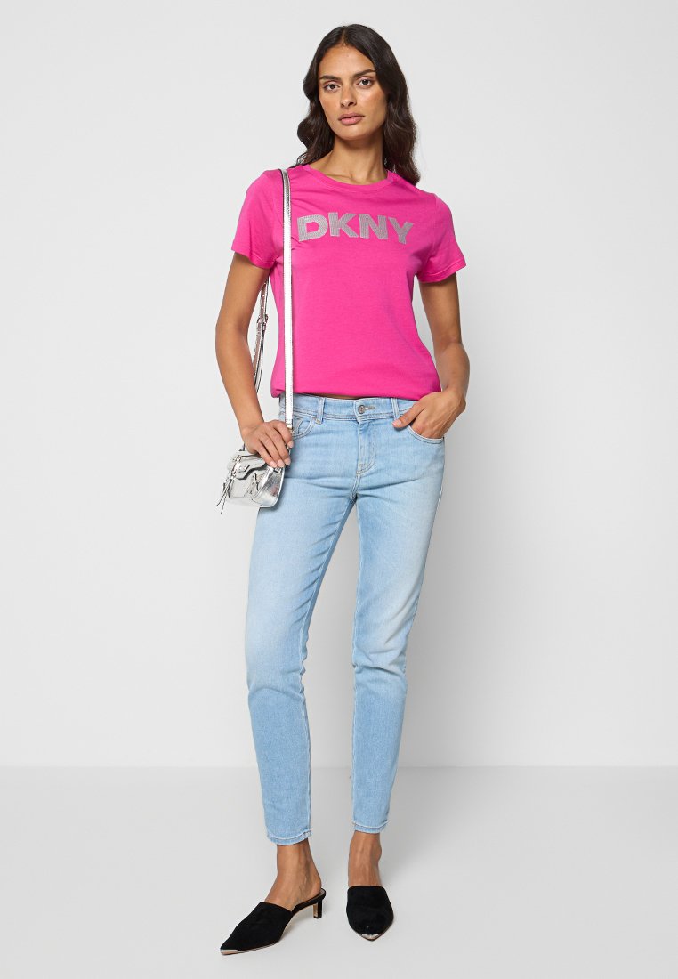 Różowa koszulka DKNY z srebrnym logo, w połączeniu z jasnoniebieskimi rurkami. Modelka nosi czarne mule i małą srebrną torebkę na ramię.