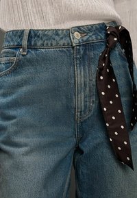 Modré riflové nohavice s vysokým pásom a čiernou mašľou s bodkami pripnutou na pútku opasku, zobrazujúce opotrebovaný vzhľad a bežné švy.