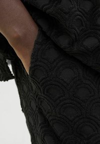 Black textured fabric featuring a raised scallop pattern, with soft, tactile fibers visible at the edges. Close-up of the textile detail.