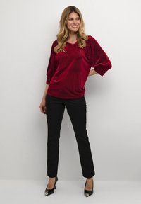 Top in velluto rosso con righe verticali, taglio rilassato e maniche a sbuffo. Abbinato a pantaloni neri slim e tacchi alti neri.