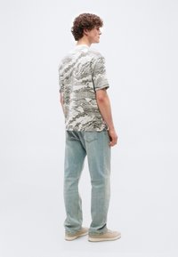 Νέος άνδρας με σγουρά μαλλιά που στέκεται, φοράει γκρι-λευκό πουκάμισο με κοντά μανίκια και σχέδιο, ανοιχτόχρωμο μπλε τζιν και μπεζ πάνινα παπούτσια.