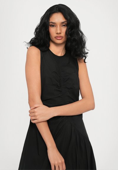 Black sleeveless dress with ruching detail at the front, flowing skirt, and soft texture. Model displays folded arms against a plain background.