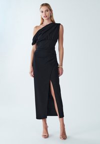 Abito nero con design a spalla unica, corpino arricciato e uno spacco laterale. Realizzato in tessuto liscio, presenta una silhouette aderente e lunghezza fino al pavimento.