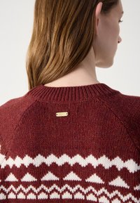 Lila testű kötött pulóver, fehér geometrikus mintákkal, bordás nyakkal és egy kis arany logólemezzel a hátán. Texturált felület.