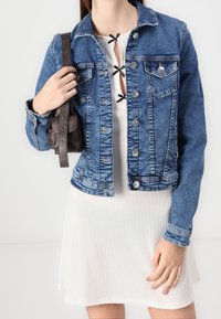 Denim jacket in medium blue with a fitted design, button closure, two chest pockets, and subtle fading, paired with a white ribbed dress.
