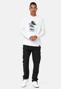 Camicia bianca a maniche lunghe con una grafica colorata di Mickey Mouse, pantaloni cargo neri e sneakers bianche. Stile streetwear semplice e informale.