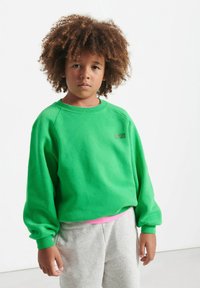 Felpa in pile verde con colletto rotondo e polsini a costine, abbinata a pantaloni grigi. Presenta un piccolo logo testuale sul petto.