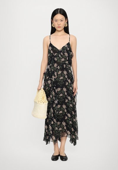 Robe maxi noire avec motif floral, détails en volants, bretelles réglables et ourlet fluide. Le modèle tient un sac tissé, de couleur jaune clair.