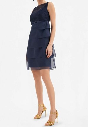 Frau trägt ein ärmelloses, marineblaues, geschichtetes Kleid mit Spitzenoberteil und goldenen Highheel-Sandalen, steht vor weißem Hintergrund.