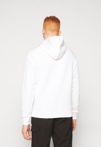 Un sweat à capuche blanc avec des cordons, des poignets et un ourlet côtelés, fait en tissu doux. Porté par-dessus un pantalon noir, vu de derrière.