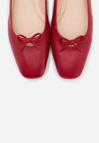 Ballerines en cuir rouge avec un talon carré, ornées d'un nœud à l'avant et d'une texture lisse. À l'intérieur, une doublure nude.