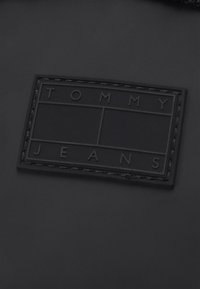 Etichetta rettangolare nera con testo in rilievo "TOMMY JEANS," circondata da un sottile bordo e cuciture delineate, su uno sfondo nero opaco.