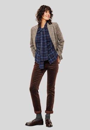 Femme debout avec les mains dans les poches, portant un blazer à carreaux, un chemisier à pois bleu avec un nœud, un pantalon brun, des chaussettes grises et des mocassins marron foncé.