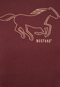 Camiseta burdeos con un contorno dorado de un caballo corriendo y la palabra "MUSTANG" en texto negrita debajo. Textura suave, diseño informal.