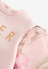 Hellrosa Sweatshirt mit goldener Schrift und floralem Druck, mit transparenten Rüscheärmeln. Weicher Stoff, lässiges Design und feminine Akzente.