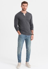 Man som bär en mörkgrå långärmad henley-tröja, ljusblå jeans, beige sneakers och står med en hand i fickan mot en vit bakgrund.